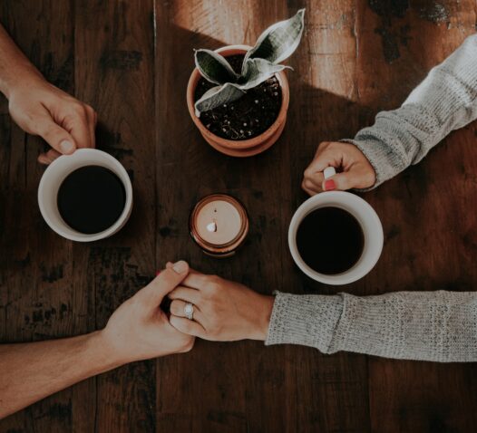 Två koppar kaffe och en grön växt står på ett bord medan två personer håller varandra i handen.